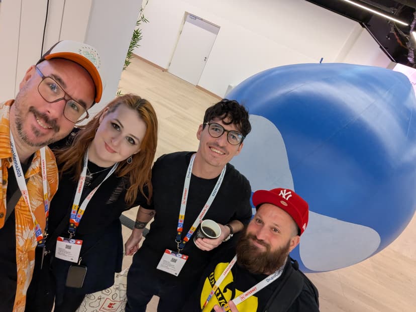 A woman and three men pose for a selfie in front of a large blue inflatable Drupal mascot in a large brightly lit room at DrupalCon 2025